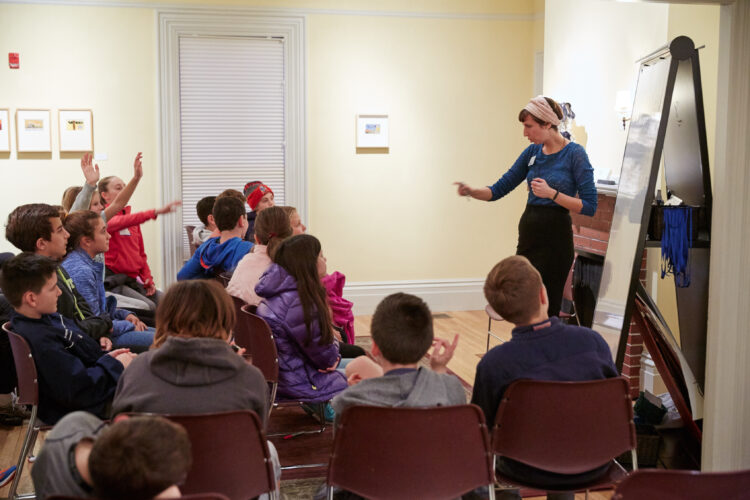 An adult woman stands in front of a white board teaching a group of middle-school-aged students. Several students have their hands raised and the adult teacher is pointing her hand to call on one of them.